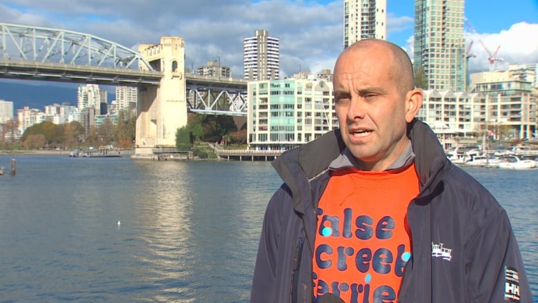 A bald man speaks in the foreground of a large water body and buildings behind him.