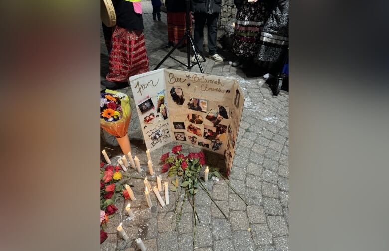 Roses lay on the ground, while candles are light up in front a board with pictures.