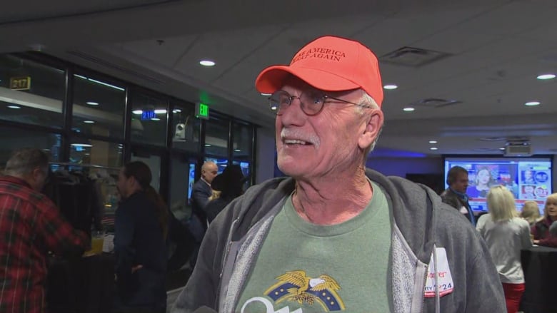 A man in a green t-shirt, grey sweater and orange hat smiles as he stands in a room with other people.