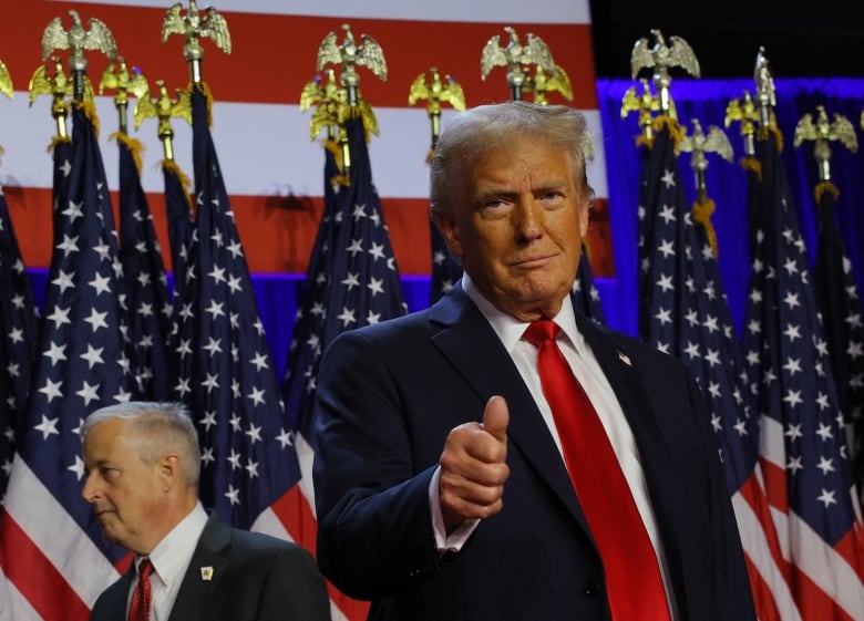 A man in a suit with a red tie gives a thumbs up on a stage decorated with American flags.