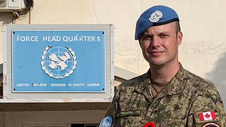 A man in military uniform with a Canadian flag on the arm and a blue UN beret stands outside a UN headquarters on a sunny day in South Sudan.