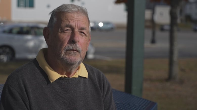 An older man with short grey in a brown sweater sits squinting into the camera.