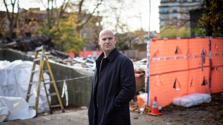 A bald man with a navy blue coat stands in front of a demolished building. 