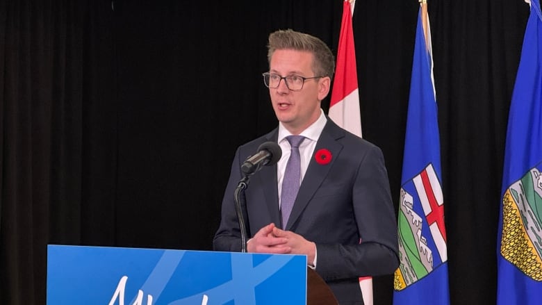 Nate Glubish stands at a lectern with the blue Alberta logo on the front. He is wearing a dark grey suit, light coloured shirt and purple tie and has a poppy in his lapel. Black curtains and Alberta and Canada flags are in the background.