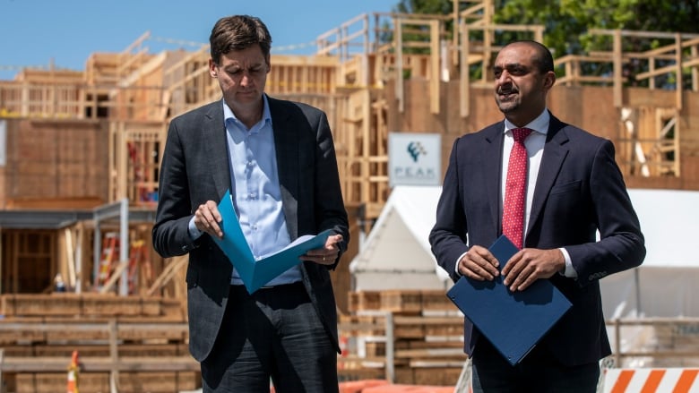 Two tall men are pictured with files in front of an under-construction home.