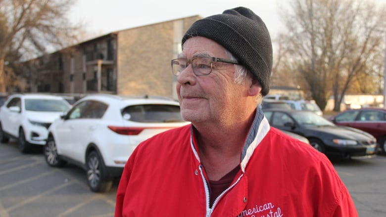 A man in a black toque and a red and white jacket stands in a parking lot.