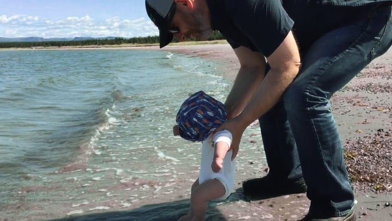 Man holding a toddler on the beach, letting toddler get his feet wet.