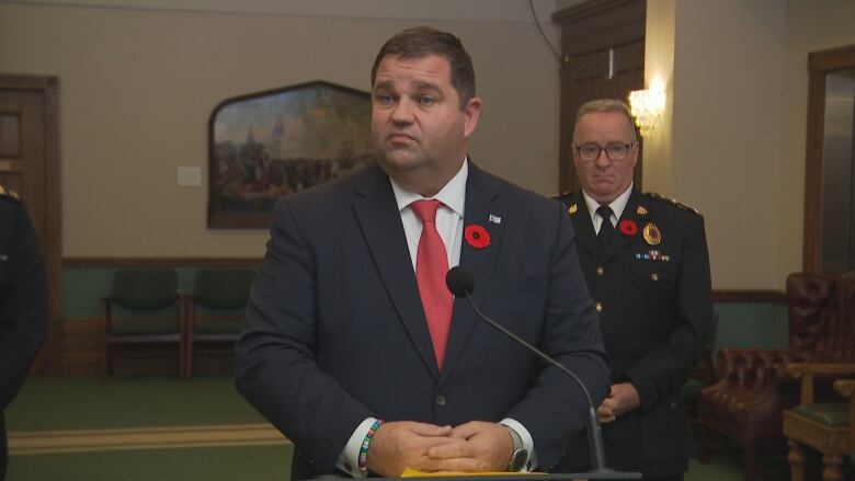 A man in a suit and red tie, with a police officer in uniform behind him.