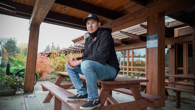 A man wearing jeans and a black jacket sits on a picnic table.