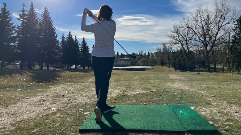 A woman golfing in a white shirt has just finished her swing. She is standing on a green golf mat and looking out towards the fairway. 