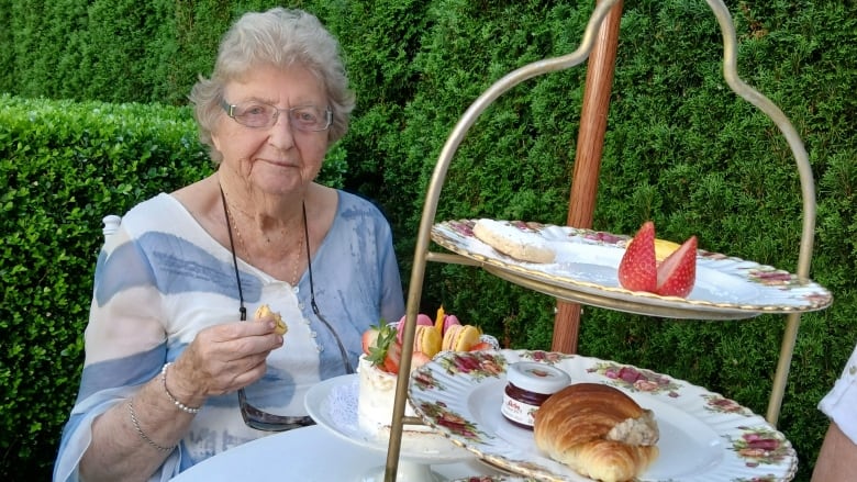 A woman with grey hair and glasses sits at a table covered in fine china and sweets