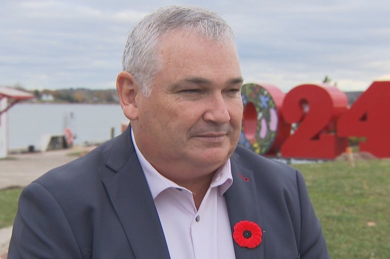 Man in dark blue suit, open white shirt, red oppy, stands in front of a large sign in background that says 2024. 