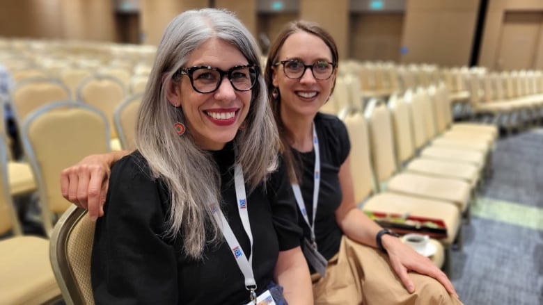 Michelle Scott has long grey hair and glasses. She's wearing a black shirt and sitting in a lecture theatre alongside Heather Bensler, who has long brown hair and glasses.