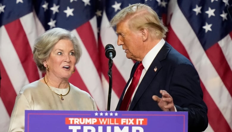 A man and a woman stand behind a podium that has a microphone. There are American flags in the background. 