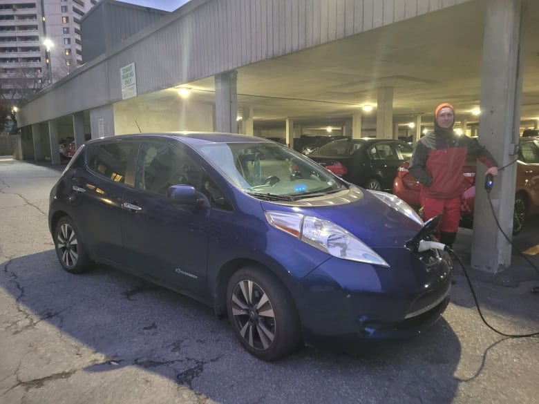 A man wearing wind protective hear stands next to a blue car plugged into an outdoor outlet.