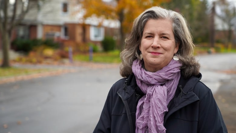 A woman with with a purple scarf and black jacket stands on a tree-lined street