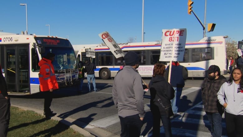 Striking employees standing with signs at a picket line as two buses drive by