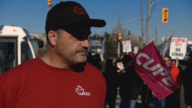 A man wearing a red CUPE shirt standing in front of workers at the picket line wearing a hat