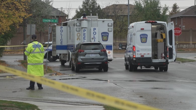 Two police vans that say Peel Police are seen behind yellow police tape. An officer in a yellow coat stands nearby. 