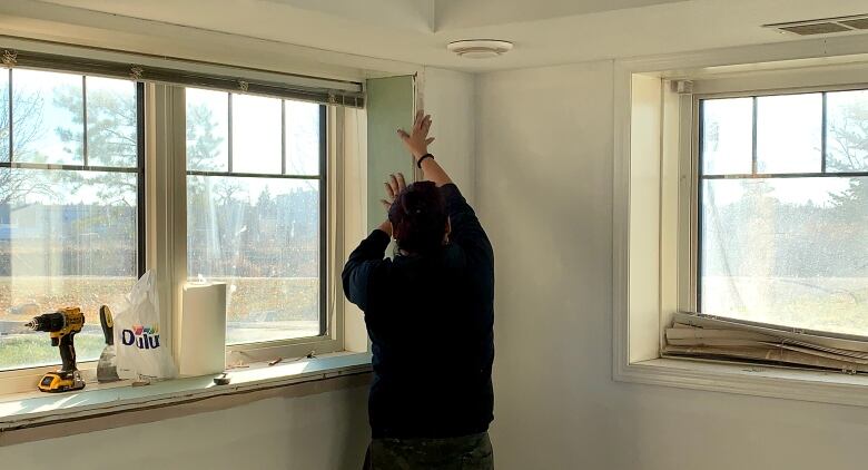 A woman holds a piece of drywall against a window ledge in an apartment suite.