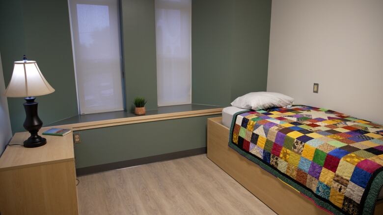 A bedroom with green walls, a nightside table with a lamp on it, and a single bed with a colourful quilt.