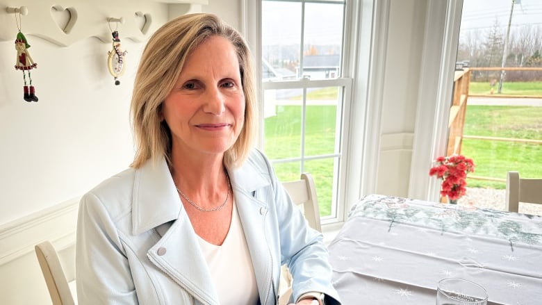 A woman with blonde hair wearing a light blue blazer and white shirt clasps her hands at a kitchen table and smiles.