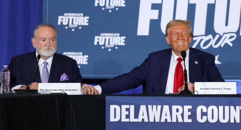 Two men in suits sit at a table on a stage.