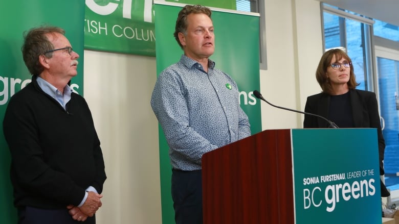 A man speaks while in front of a green banner, while a man and a woman flank him.