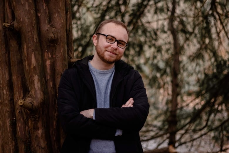 Man with glasses smiling and leaning against a tree. He's wearing a grey shirt and a black jacket.