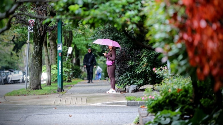 A woman with a pink umbrella stand on the sidewalk