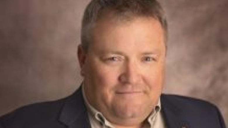 A professional head and upper body portrait of a man in about his 40s with light coloured short hair wearing a dark blue suit and light coloured button-down collared shirt with the top button open.