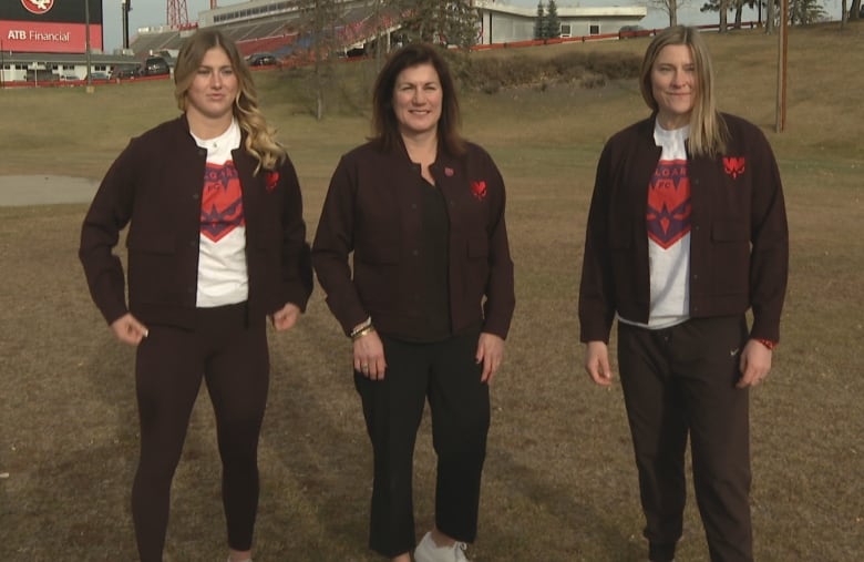 Three women with black jackets on stand in a field.