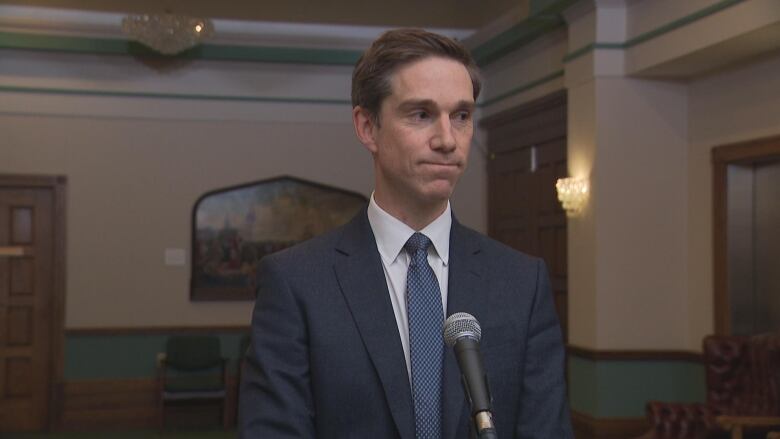 N.L. Health Minister John Hogan stands behind a microphone. He is wearing a navy blue blazer.