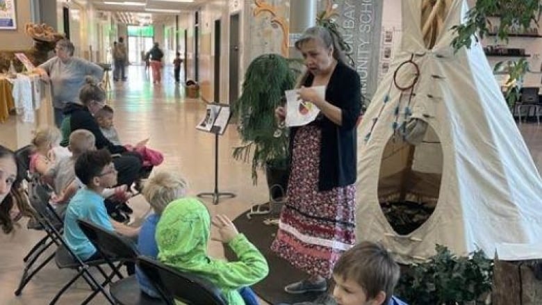 A women, wearing a long skirt and hair half hair, stand in front of a tipi while holding a book to read to children in school.