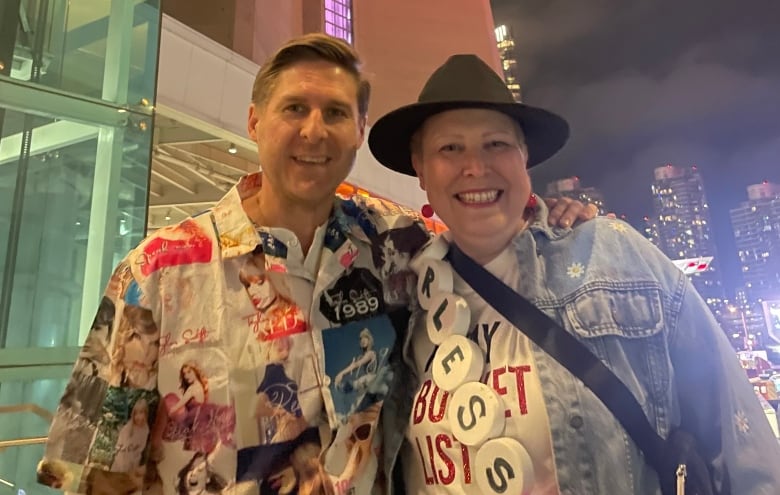 Man in shirt covered in photos of Taylor Swift poses with woman in black brimmed hat.