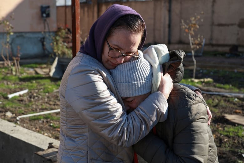 A woman in glasses and wearing a hoodie hugs another woman who has a toque and a winter coat on, in an outdoor photo.