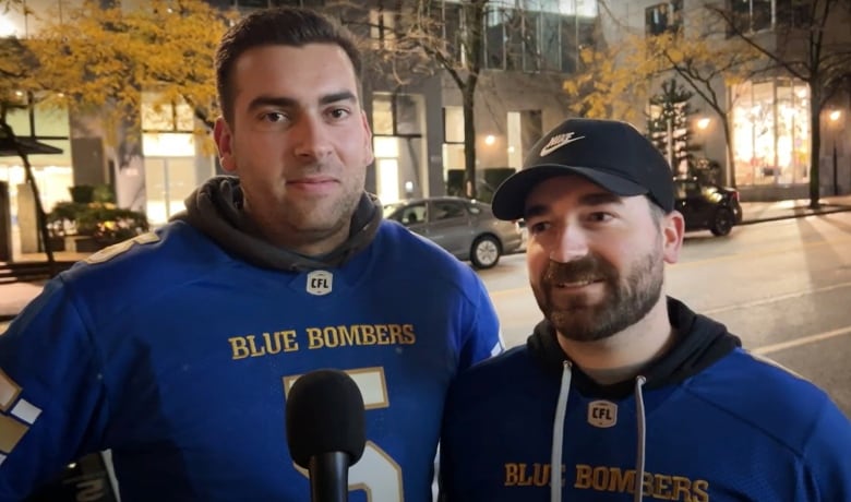 Two men in blue jerseys face a camera and speak into a microphone while outside on a street.