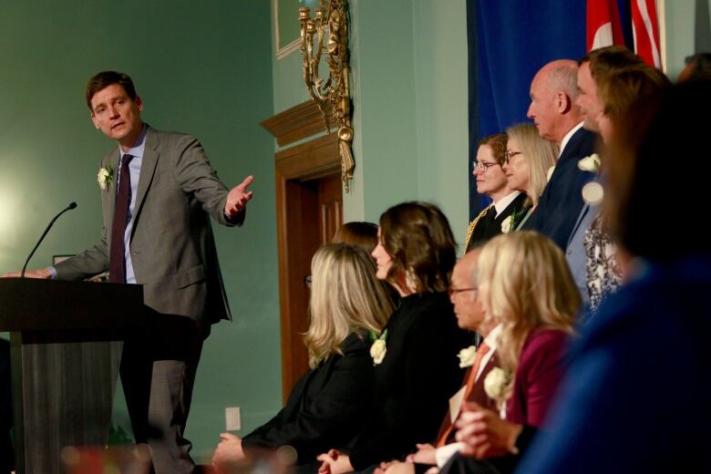 A man stands at a podium and gestures toward a group of people on stage next to him