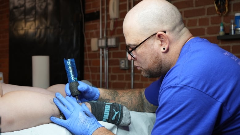 A bald man wearing glasses hold a tattoo gun as he is tattooing a person's calf leg.