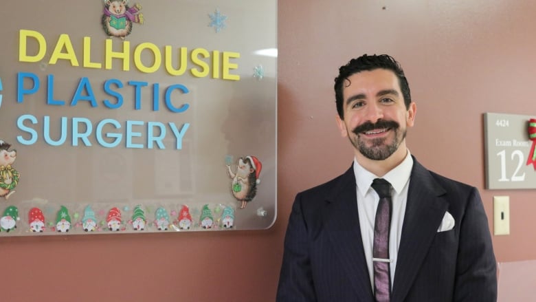 A smiling man with slicked back hair, a mustache and a beard wears a suit and a tie. He stands next to a sign saying 