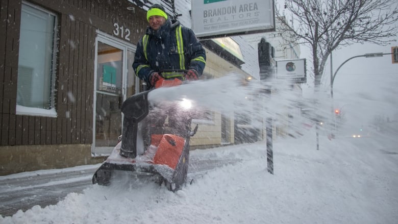 A man in a snowsuit uses a snowblower to clear a sidewalk