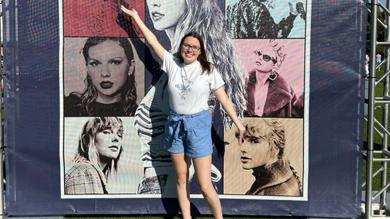 A smiling woman in shorts and a t-shirt standing in front of a giant Taylor Swift poster.