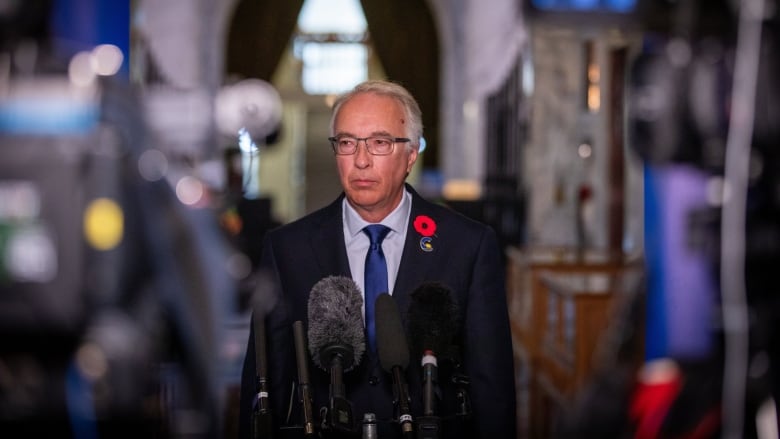 A man looks to his right as he stands in front of a gaggle of microphones.