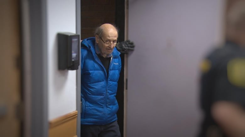 A man in a blue coat walking through a doorway with his hands behind his back.