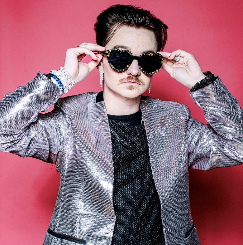 A male adjusts his dark shades during a portrait, wearing a glittery silver collarless jacket as well as earrings and friendship bracelets.