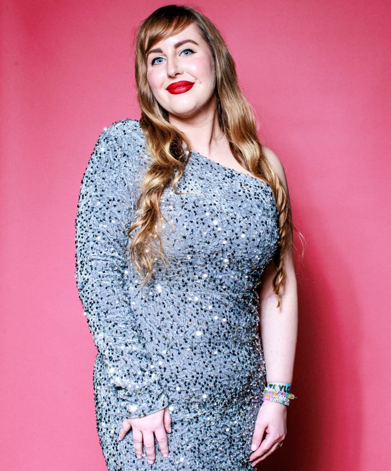 A woman with long blond hair and red lips, in a sequin gown with one shoulder bare, smiles for the camera.