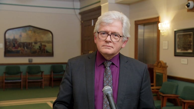 Man with grey hair in dark grey suit with purple button up shirt.