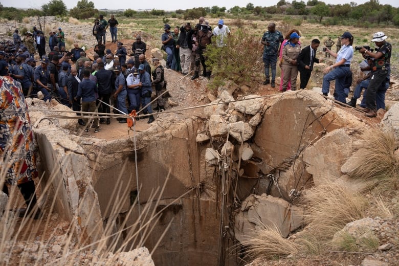 Dozens of people, including uniformed police officers and men in suits, stand around the edge of a rocky mine shaft.