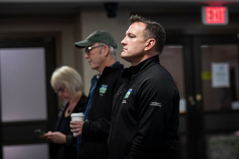 A man with a stern look on his face seen in profile stands next to another man and a woman looking at someone in front of them off camera.