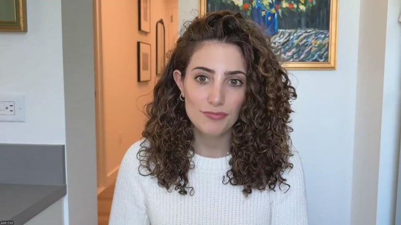 A woman sits in a room, with a painting behind her.